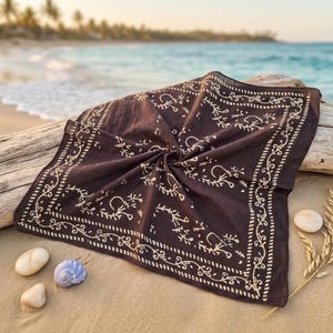 K&ouml;nnte beinhalten: Ein dunkelbraunes Bandana mit cremefarbenem Blumenmuster, das auf Treibholz am Strand liegt. Das quadratische Tuch hat einen detaillierten Rand. Muscheln und Weizen&auml;hren befinden sich in der N&auml;he, mit dem Meer im Hintergrund.