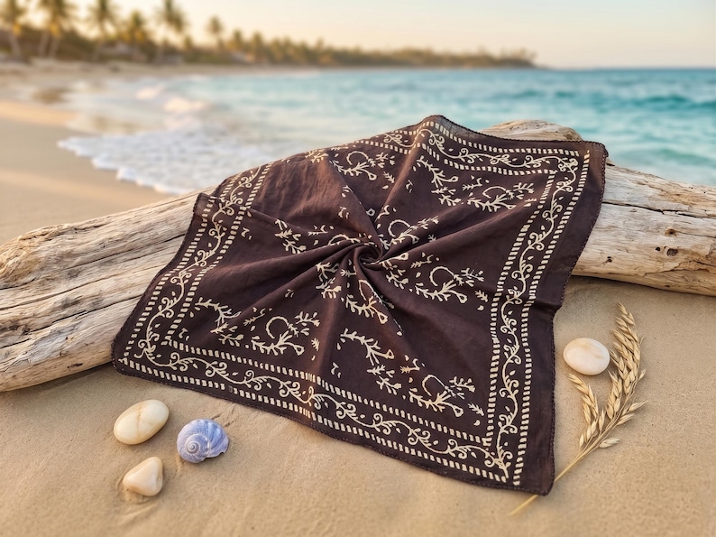K&ouml;nnte beinhalten: Ein dunkelbraunes Bandana mit cremefarbenem Blumenmuster, das auf Treibholz am Strand liegt. Das quadratische Tuch hat einen detaillierten Rand. Muscheln und Weizen&auml;hren befinden sich in der N&auml;he, mit dem Meer im Hintergrund.