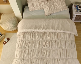 May include: A beige bed set featuring a ruched comforter and two matching pillowcases. The bedding is arranged on a bed with a light green sheet. The room has a neutral color palette with a rug and a small side table.