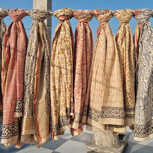 May include: A collection of patterned scarves in various colors, including cream, red, and tan, hang from a metal bar against a blue sky. The scarves feature block print designs with geometric and floral motifs, showcasing a range of patterns and styles.
