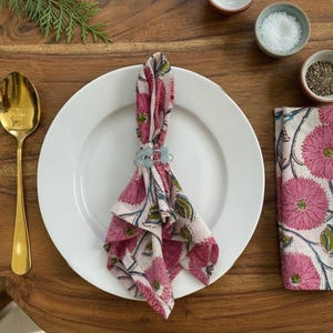 May include: A dining table setting featuring a white plate, gold cutlery, and a folded napkin secured by a light blue napkin ring. The napkin has a pink floral design. Small bowls of salt and pepper are also visible.
