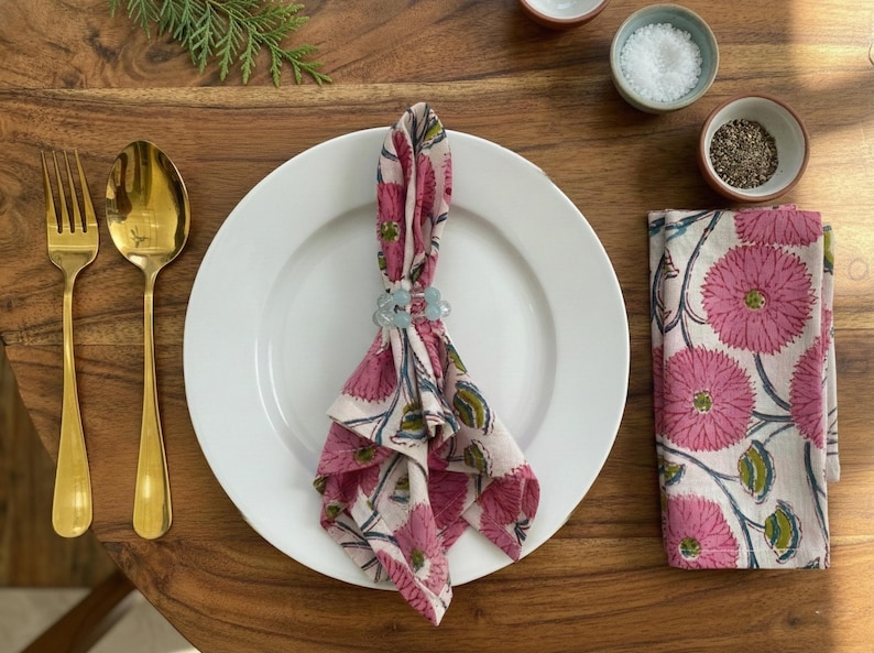 May include: A dining table setting featuring a white plate, gold cutlery, and a folded napkin secured by a light blue napkin ring. The napkin has a pink floral design. Small bowls of salt and pepper are also visible.