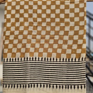 May include: A rectangular textile with a repeating pattern of brown and white squares. Below the squares are horizontal black stripes and a border of black teardrop shapes. The fabric is hanging from a wooden rod.
