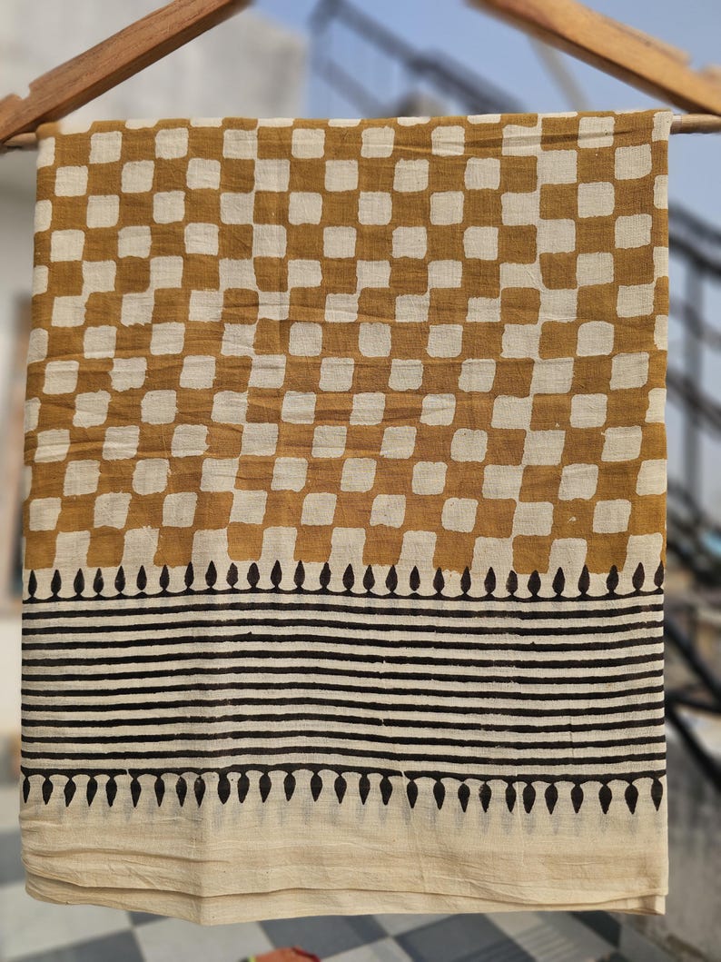 May include: A rectangular textile with a repeating pattern of brown and white squares. Below the squares are horizontal black stripes and a border of black teardrop shapes. The fabric is hanging from a wooden rod.