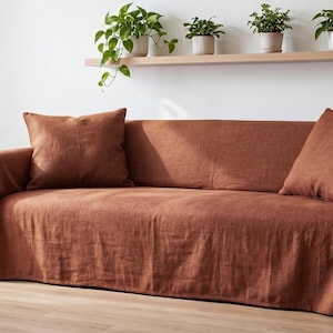 Può includere: Un copridivano color ruggine drappeggiato su un divano, caratterizzato da una finitura in tessuto testurizzato. La copertura include due cuscini quadrati abbinati posizionati sulla seduta, con il tessuto che scende sui braccioli e sui lati.
