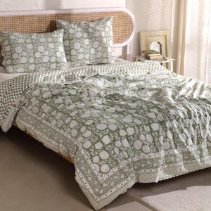 May include: A bed with a green and white floral patterned quilt and two matching pillowcases. The bedding has a detailed border design and is set on a bed with a woven headboard. The color palette is soft and inviting.