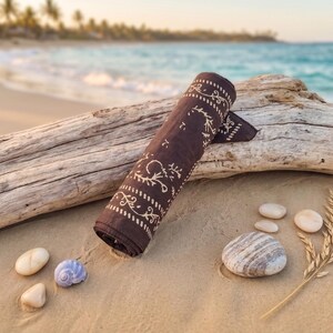 K&ouml;nnte beinhalten: Gerolltes braunes Bandana mit wei&szlig;em floralem Muster, das auf einem St&uuml;ck Treibholz an einem Sandstrand liegt. Mehrere kleine Steine und eine Muschel sind verstreut. Das Meer und Palmen sind im Hintergrund zu sehen.