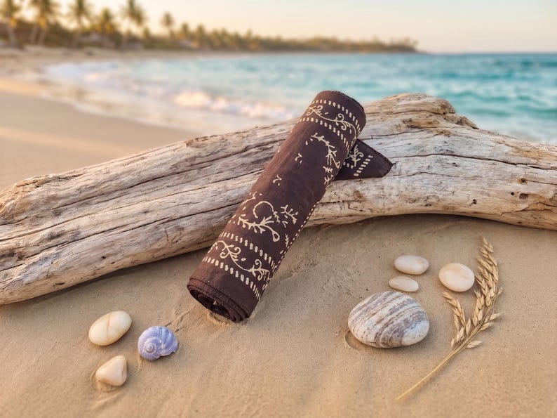 K&ouml;nnte beinhalten: Gerolltes braunes Bandana mit wei&szlig;em floralem Muster, das auf einem St&uuml;ck Treibholz an einem Sandstrand liegt. Mehrere kleine Steine und eine Muschel sind verstreut. Das Meer und Palmen sind im Hintergrund zu sehen.