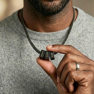 Puede incluir: Un collar de cadena negra con cuatro colgantes rectangulares. Cada colgante tiene grabada la palabra "Forever", "Loved" y "I Am". El collar lo lleva una persona con un jersey gris.