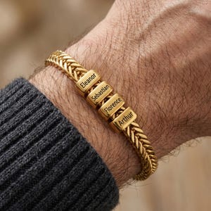 May include: Gold-colored bracelet with a woven chain design. The bracelet features four rectangular gold beads, each engraved with a name: Eleanor, Sebastian, Florence, and Arthur. The bracelet is shown on a person's wrist.