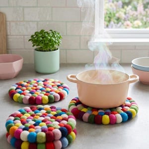 May include: A peach-coloured cooking pot with steam rising from it sits on a colourful, round trivet made of felt balls. Two additional trivets are visible, along with a small potted plant and bowls, all on a light-coloured countertop.