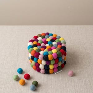 May include: A circular trivet constructed from numerous small, multicolored felt balls arranged in a dense, layered pattern. The colors include red, orange, yellow, green, blue, purple, pink, white, and black. Several loose felt balls lie nearby.