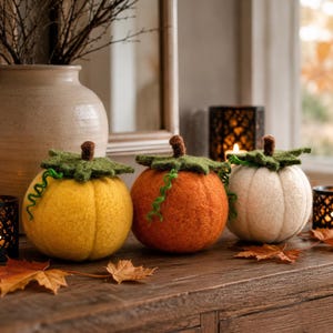 May include: Three round needle-felted pumpkins in yellow, orange, and white. Each pumpkin features a textured green felt leaf topper, a small brown stem, and a thin green curly vine accent. The pumpkins have a soft, fuzzy, wool-like appearance.