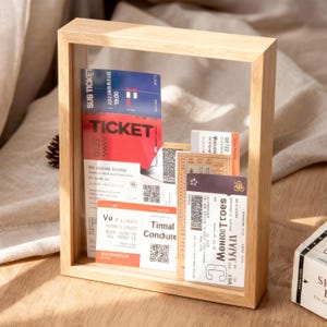 Può includere: Una cornice espositiva in legno con un fronte in vetro trasparente, contenente vari biglietti e carte. I biglietti sono rossi, blu e beige, con testo stampato e codici QR. La cornice è in legno chiaro.