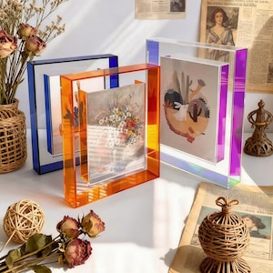 May include: Three acrylic picture frames in blue, orange, and iridescent purple. Each frame holds a different image, including a floral still life and a desert scene with a rabbit. The frames are rectangular with a clear center.