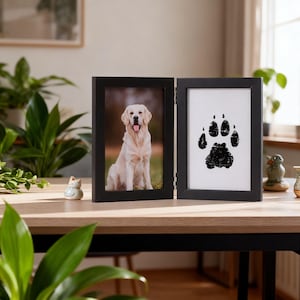May include: A black, hinged picture frame with two panels. One panel shows a photo of a golden retriever dog. The other panel displays a black paw print on a white background. The frame is on a wooden table.