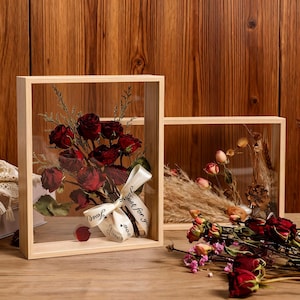 May include: Two wooden framed shadow boxes with glass fronts. One box contains a bouquet of dried red roses tied with a ribbon that reads "Just for you". The other box contains dried flowers and grasses. The background is a wooden wall.