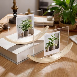 May include: Two clear acrylic photo frames with a wooden base, each holding a photo of a potted plant. The frames have a curved, rocking-style base. The photos show green plants in gray pots.
