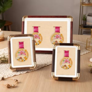 Puede incluir: Tres medallas enmarcadas con bordes dorados, cada una con un diseño de flor rosa y una cinta roja. Los marcos tienen detalles dorados, marrones y blancos. Las medallas se exhiben sobre un fondo beige.