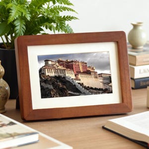 May include: A rectangular wooden picture frame with a photograph of a large building complex on a hillside. The frame is a warm brown color, and the photograph has a white border. The building is a mix of white and red.
