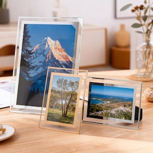 Puede incluir: Tres marcos de fotos de vidrio transparente de diferentes tamaños, cada uno con una fotografía de paisaje diferente. Los marcos se exhiben sobre una superficie de madera clara. Un marco tiene un borde dorado.