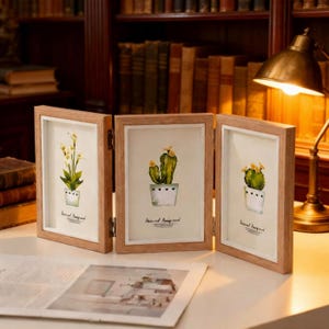 Puede incluir: Un marco de fotos de madera de tres paneles con estampados de acuarela botánicos. Los estampados presentan plantas en macetas, incluyendo cactus y flores, con el texto "Natural Background". El marco está abierto sobre una superficie blanca.