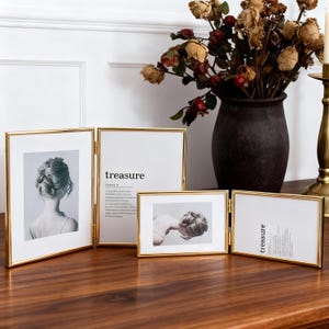 May include: A collection of gold-framed photo displays, some with images of a woman's back and the word "treasure" printed on white paper. The frames are arranged on a wooden surface, with a vase of dried flowers in the background.