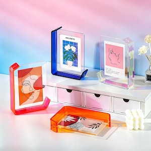 Puede incluir: Marcos de fotos acrílicos en varios colores: rojo, azul y transparente. Cada marco contiene una impresión artística diferente. Un marco muestra la palabra "HOLIDAYS". Los marcos están sobre una superficie blanca con un fondo degradado rosa y azul.