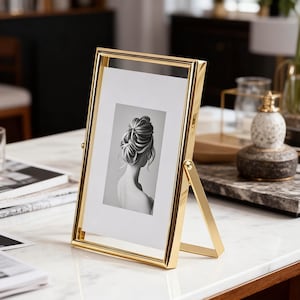 May include: A gold-colored metal picture frame with a clear glass front. The frame holds a black and white image of a woman's back and the back of her head. The frame has a hinged stand for display on a table or shelf.