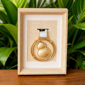 May include: A framed gold medal with a white ribbon and black and white detailing. The medal is circular with two connected smaller circles. The frame is light brown wood, and the background is a light tan colour.