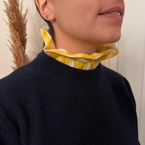 Puede incluir: Un suéter azul marino con un cuello a cuadros amarillo y blanco. El cuello es con volantes y añade un elemento decorativo al suéter. El suéter es de estilo clásico.