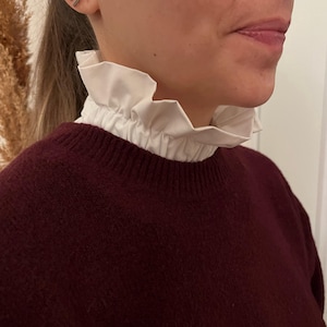 May include: A white, ruffled collar worn over a burgundy sweater. The collar is made of a crisp, white fabric and adds a touch of elegance. The sweater is a deep burgundy color, creating a contrast with the collar.