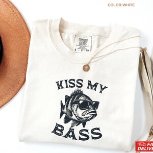 May include: White t-shirt with the phrase "KISS MY BASS" and a graphic of a fish wearing sunglasses. The shirt has a crew neck and short sleeves. A gold necklace is draped on the shirt.