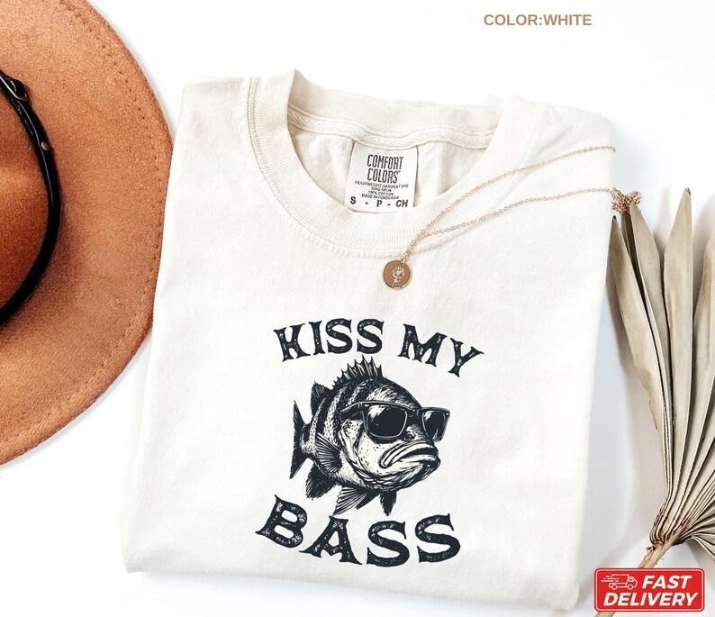 May include: White t-shirt with the phrase "KISS MY BASS" and a graphic of a fish wearing sunglasses. The shirt has a crew neck and short sleeves. A gold necklace is draped on the shirt.