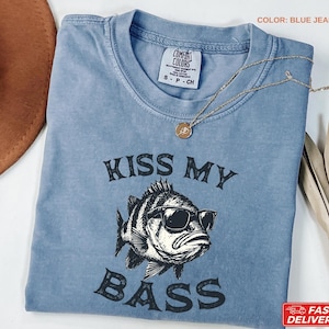 May include: Blue jean colored t-shirt with the phrase "KISS MY BASS" printed in black. The graphic features a fish wearing sunglasses. A gold necklace with a pendant is draped across the shirt.