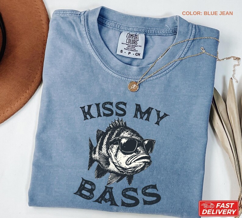 May include: Blue jean colored t-shirt with the phrase "KISS MY BASS" printed in black. The graphic features a fish wearing sunglasses. A gold necklace with a pendant is draped across the shirt.