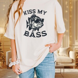 May include: Ivory-colored t-shirt with the text "KISS MY BASS" and a graphic of a fish wearing sunglasses. The shirt is a relaxed fit and has short sleeves. The color is ivory.