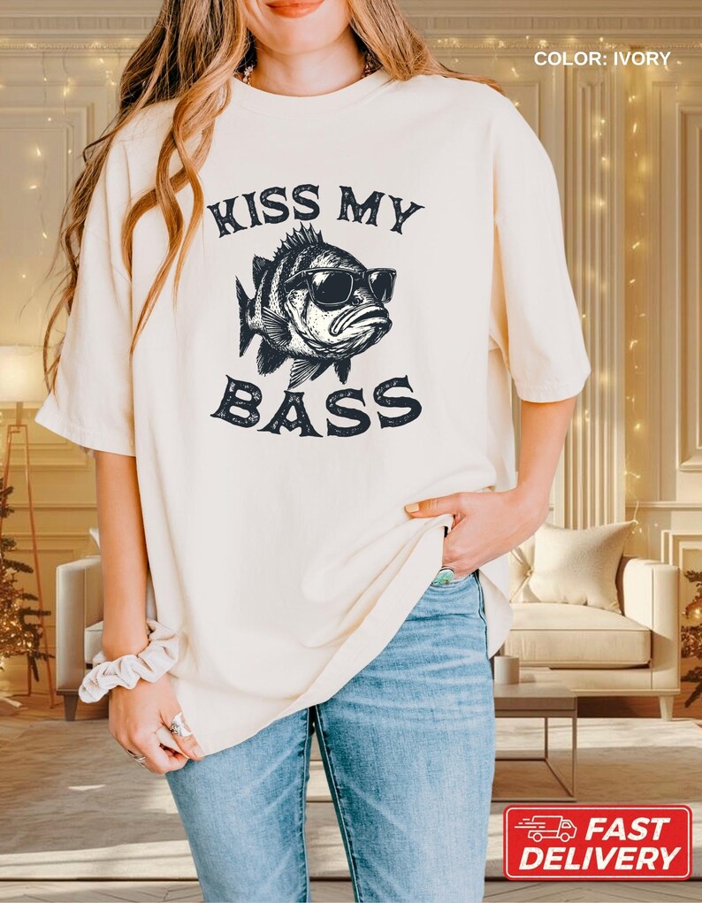 May include: Ivory-colored t-shirt with the text "KISS MY BASS" and a graphic of a fish wearing sunglasses. The shirt is a relaxed fit and has short sleeves. The color is ivory.