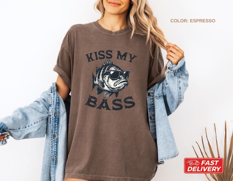 May include: Espresso-colored t-shirt with the text "KISS MY BASS" and a graphic of a fish wearing sunglasses. The shirt has short sleeves and a relaxed fit. The image also shows a denim jacket.