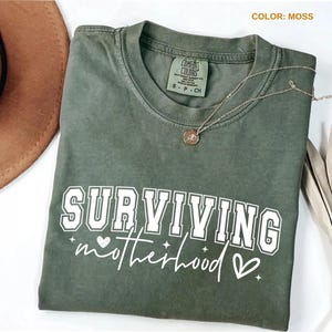 May include: A moss green Comfort Colors t-shirt with the words "SURVIVING motherhood" in white block and script lettering. A gold necklace with a pendant rests on the shirt. A brown hat is partially visible in the upper left corner.