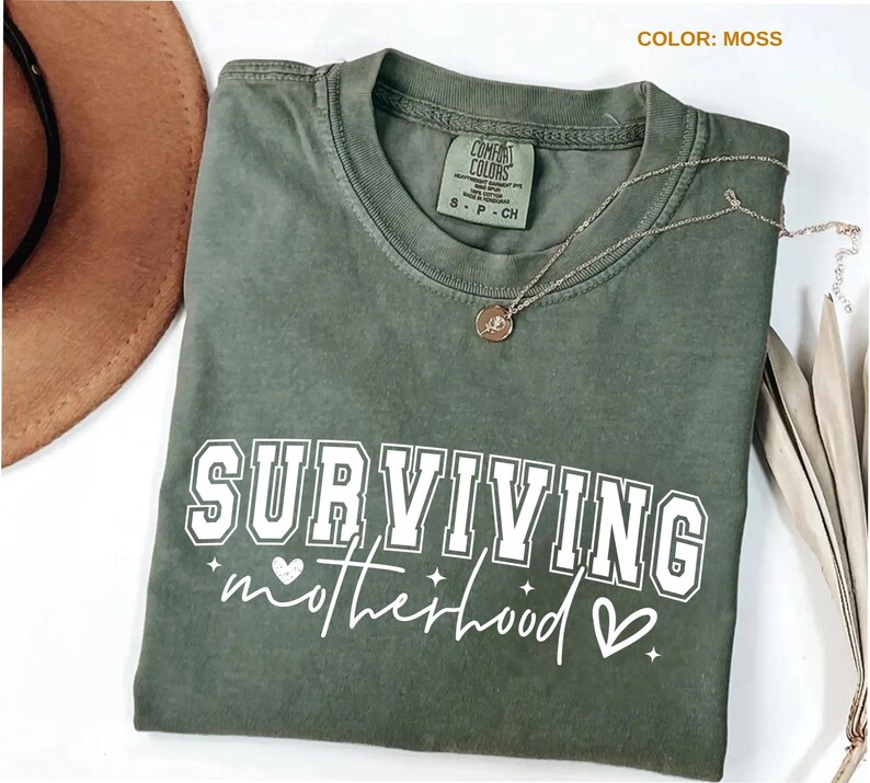 May include: A moss green Comfort Colors t-shirt with the words "SURVIVING motherhood" in white block and script lettering. A gold necklace with a pendant rests on the shirt. A brown hat is partially visible in the upper left corner.