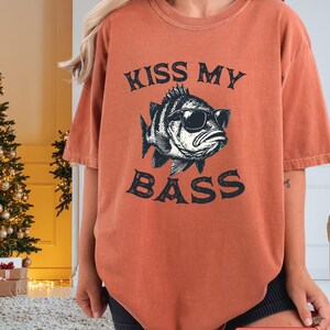 May include: A yam-colored t-shirt with the text "KISS MY BASS" above and below a graphic of a fish wearing sunglasses. The shirt has short sleeves and a relaxed fit. The image also includes a "FAST DELIVERY" sticker.