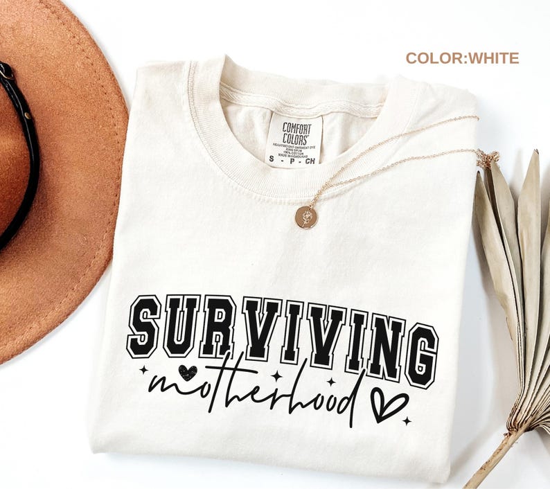 May include: Cream-colored t-shirt with the words "SURVIVING motherhood" in black lettering. The shirt has a crew neck and short sleeves. A gold necklace with a pendant is draped across the shirt. The shirt is labeled "Comfort Colors".