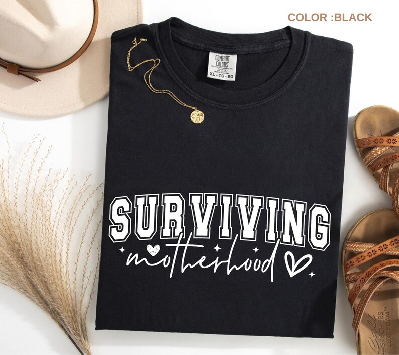 May include: Black t-shirt with the text "SURVIVING motherhood" in white block letters and script. A gold necklace with a pendant is draped on the shirt. The shirt is next to a beige hat and brown sandals.