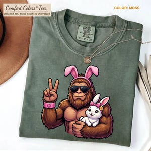 May include: Moss green Comfort Colors tee featuring a cartoon Bigfoot wearing sunglasses and pink bunny ears, holding a white bunny. The Bigfoot is giving a peace sign. The shirt has the text "Comfort Colors Tees" and "Relaxed Fit, Runs Slightly Oversized".