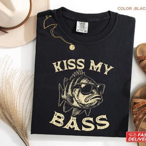 May include: Black t-shirt with the text "KISS MY BASS" and a graphic of a fish wearing sunglasses. A gold necklace is draped on the shirt. The shirt is next to a straw hat and brown sandals.