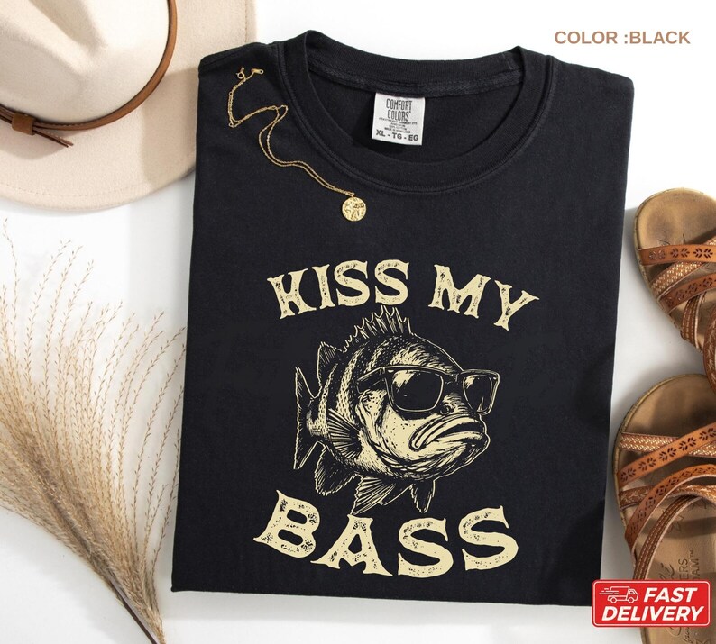 May include: Black t-shirt with the text "KISS MY BASS" and a graphic of a fish wearing sunglasses. A gold necklace is draped on the shirt. The shirt is next to a straw hat and brown sandals.
