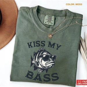 May include: A moss-colored t-shirt with the text "KISS MY BASS" and a graphic of a fish wearing sunglasses. The shirt has a crew neck and short sleeves. A gold necklace with a pendant is on the shirt.