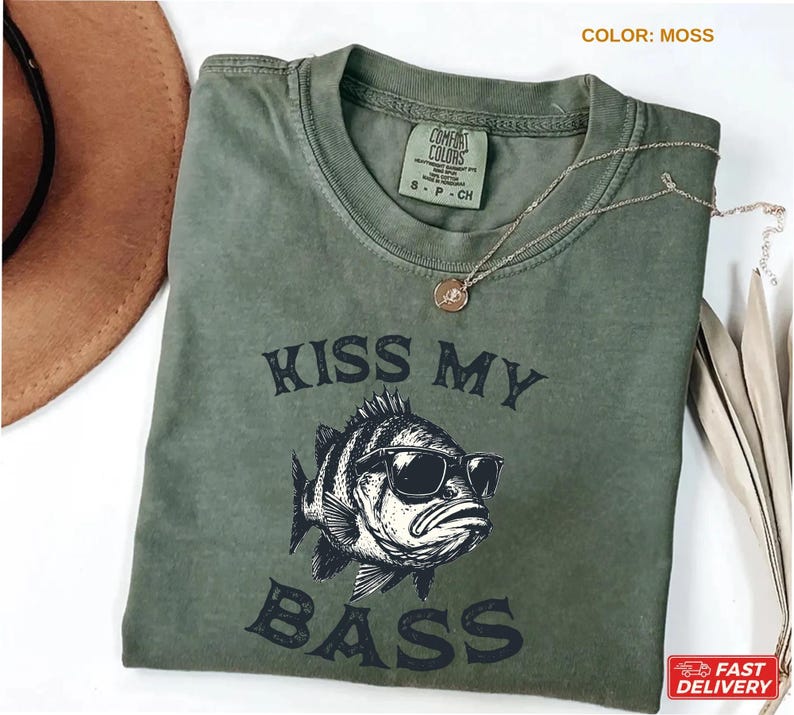 May include: A moss-colored t-shirt with the text "KISS MY BASS" and a graphic of a fish wearing sunglasses. The shirt has a crew neck and short sleeves. A gold necklace with a pendant is on the shirt.