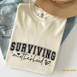 May include: Ivory-colored t-shirt with the text "SURVIVING motherhood" in black. The shirt is a crew neck and is folded on top of a pair of blue jeans. A tan hat is visible in the background.